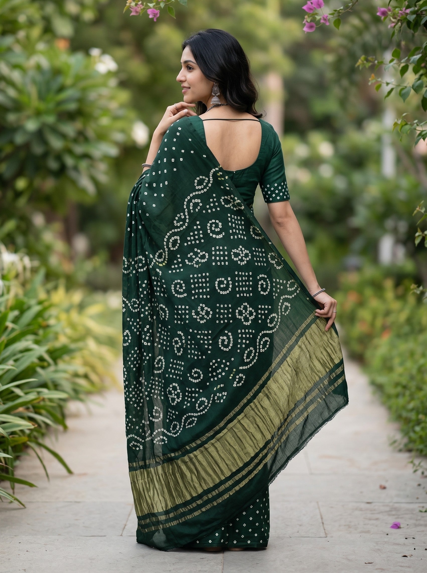 A woman wearing a dark green saree with white bandhani motifs and a gold border, standing on a garden path.