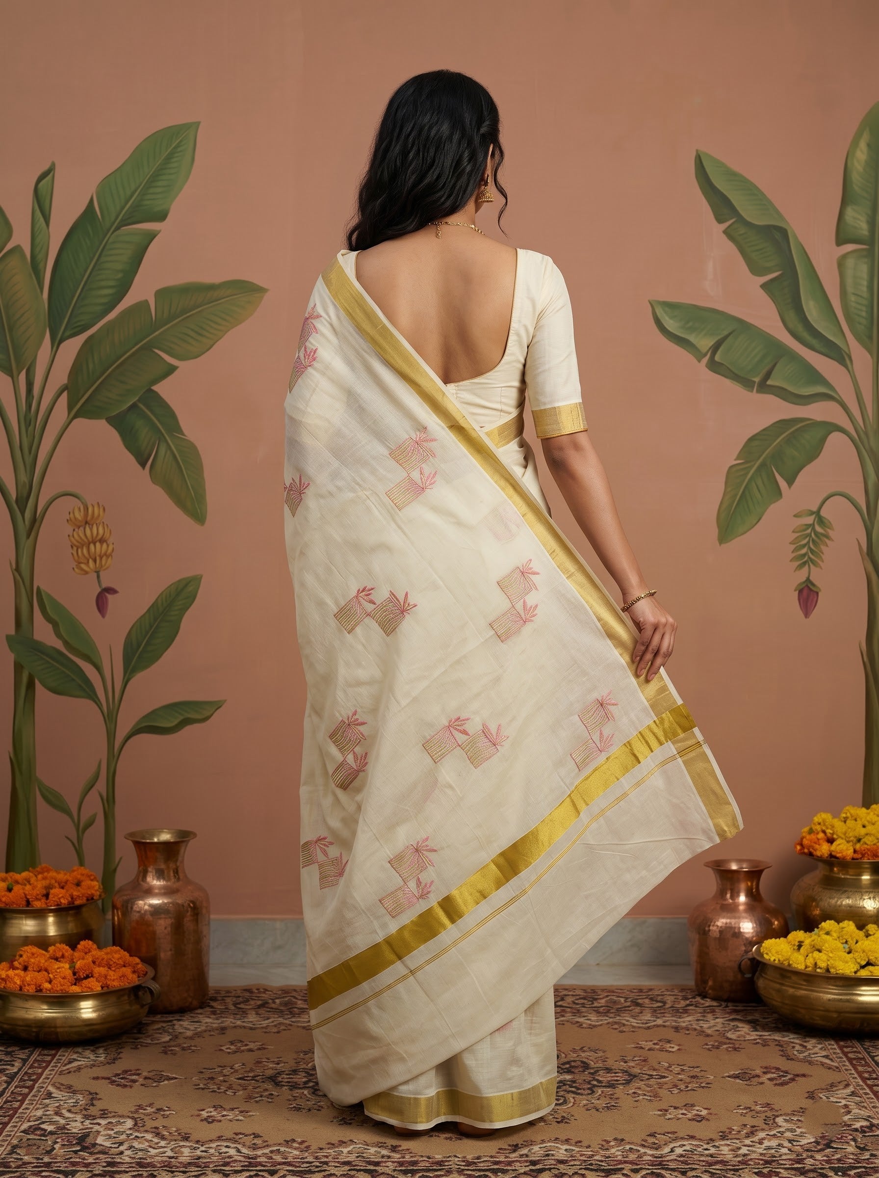 A woman stands with her back to the camera wearing a cream-colored cotton saree with pink floral motifs and gold borders, paired with a matching cream blouse. The setting features a terracotta-colored wall decorated with green banana plant illustrations, copper pots, and yellow flowers on a patterned rug.
