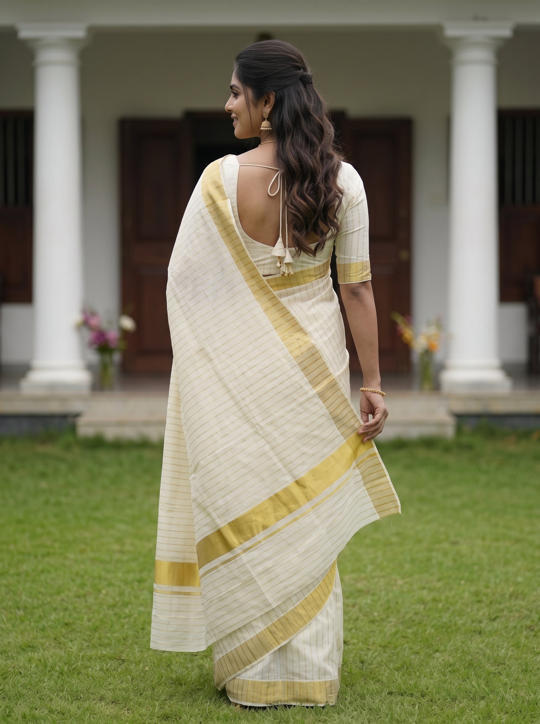 A woman wearing a cream-colored saree with gold stripes and a matching cream blouse, standing outdoors on a green lawn in front of a house with white columns.