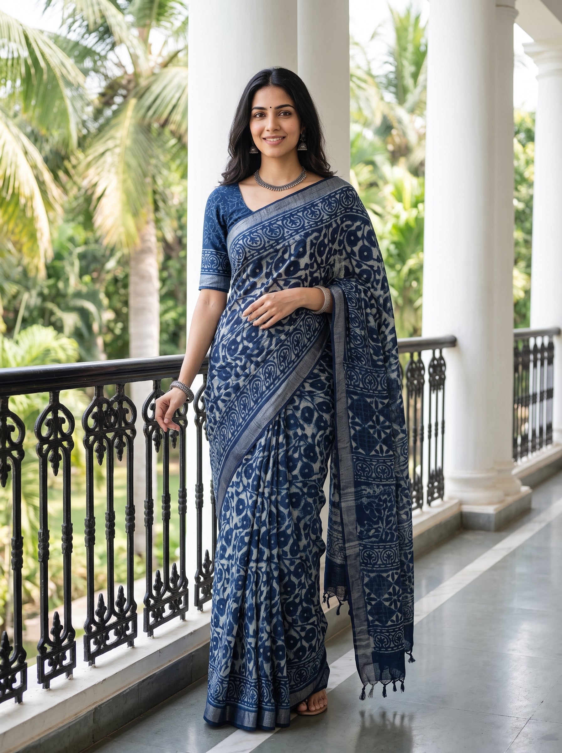 A woman wearing a navy blue and gray printed saree with a matching printed blouse, standing on a covered outdoor walkway with palm trees in the background.