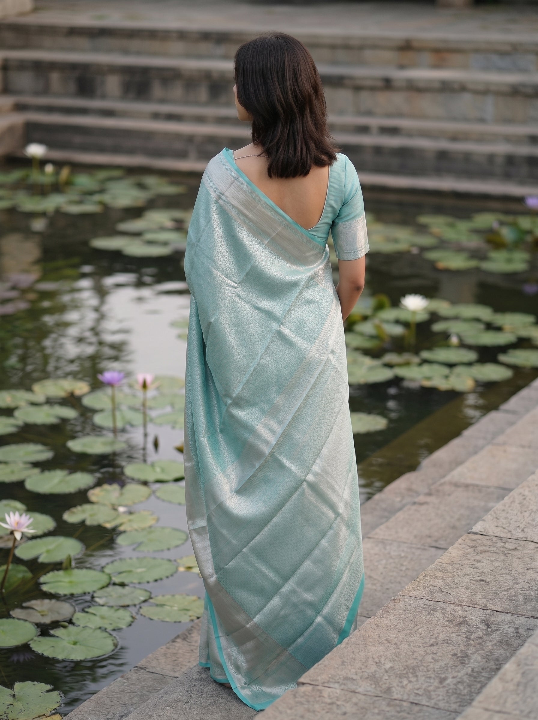 A woman viewed from behind wearing a light mint green saree with a silver border, standing on stone steps next to a pond with lily pads.
