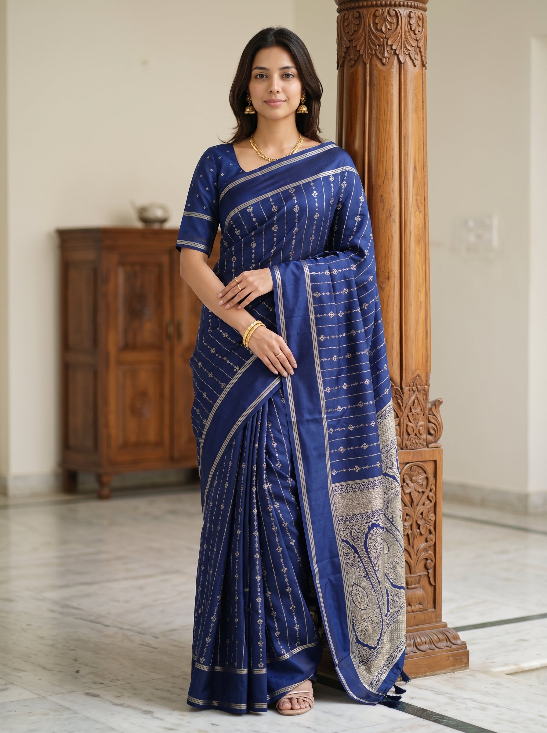 A woman wearing a royal blue cotton saree with white stripes and a silver border, standing in a traditional indoor setting next to a carved wooden pillar.