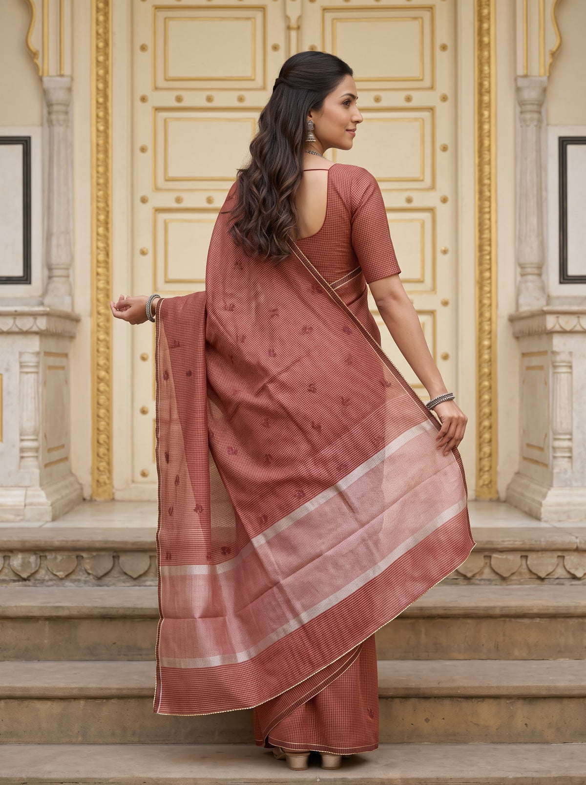 A woman wearing a dusty rose terracotta saree with a matching blouse, standing on stone steps in front of a golden-arched doorway. The saree has a subtle sheen with small motifs and a contrasting border of white and light gold stripes.