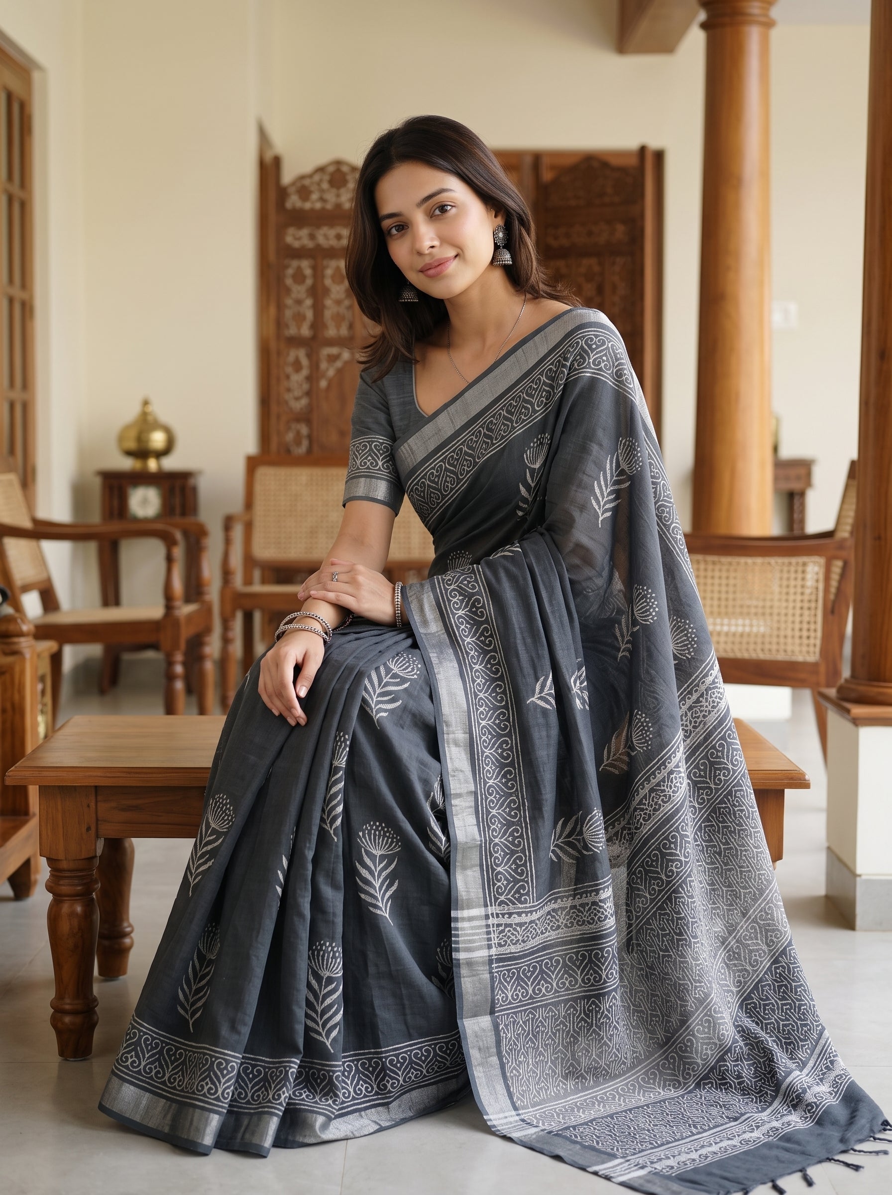 A woman wearing a dark gray saree with light gray block prints and borders, seated in a traditional indoor setting with wooden furniture.