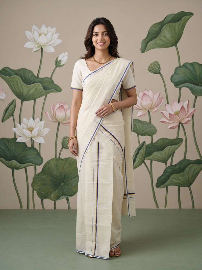 A woman modeling a cream-colored cotton saree with thin blue and green border stripes, paired with a matching cream blouse, standing in front of a painted lotus wall mural.