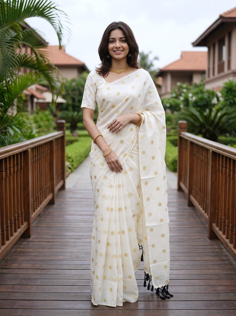 A woman wearing a white saree with gold polka dots stands on a wooden walkway in an outdoor resort setting with tropical plants and resort buildings in the background.