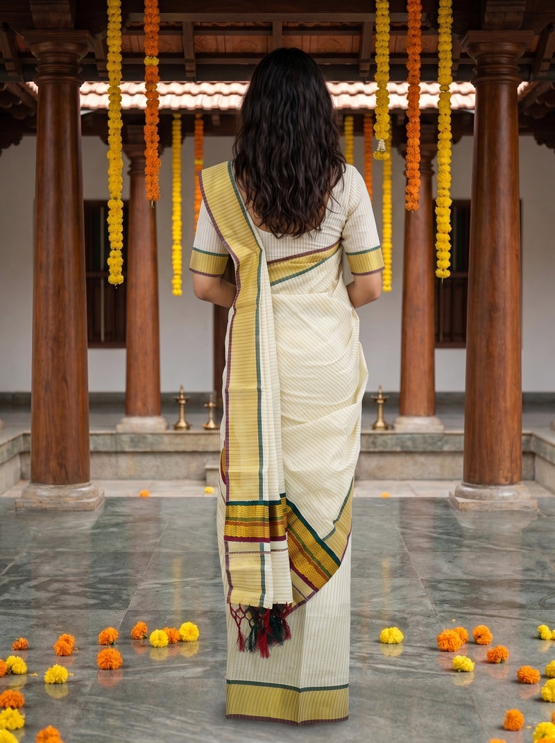 A woman viewed from behind wearing a cream-colored cotton saree with a yellow, green, and red border, standing in a traditional courtyard with wooden pillars and hanging marigold garlands.