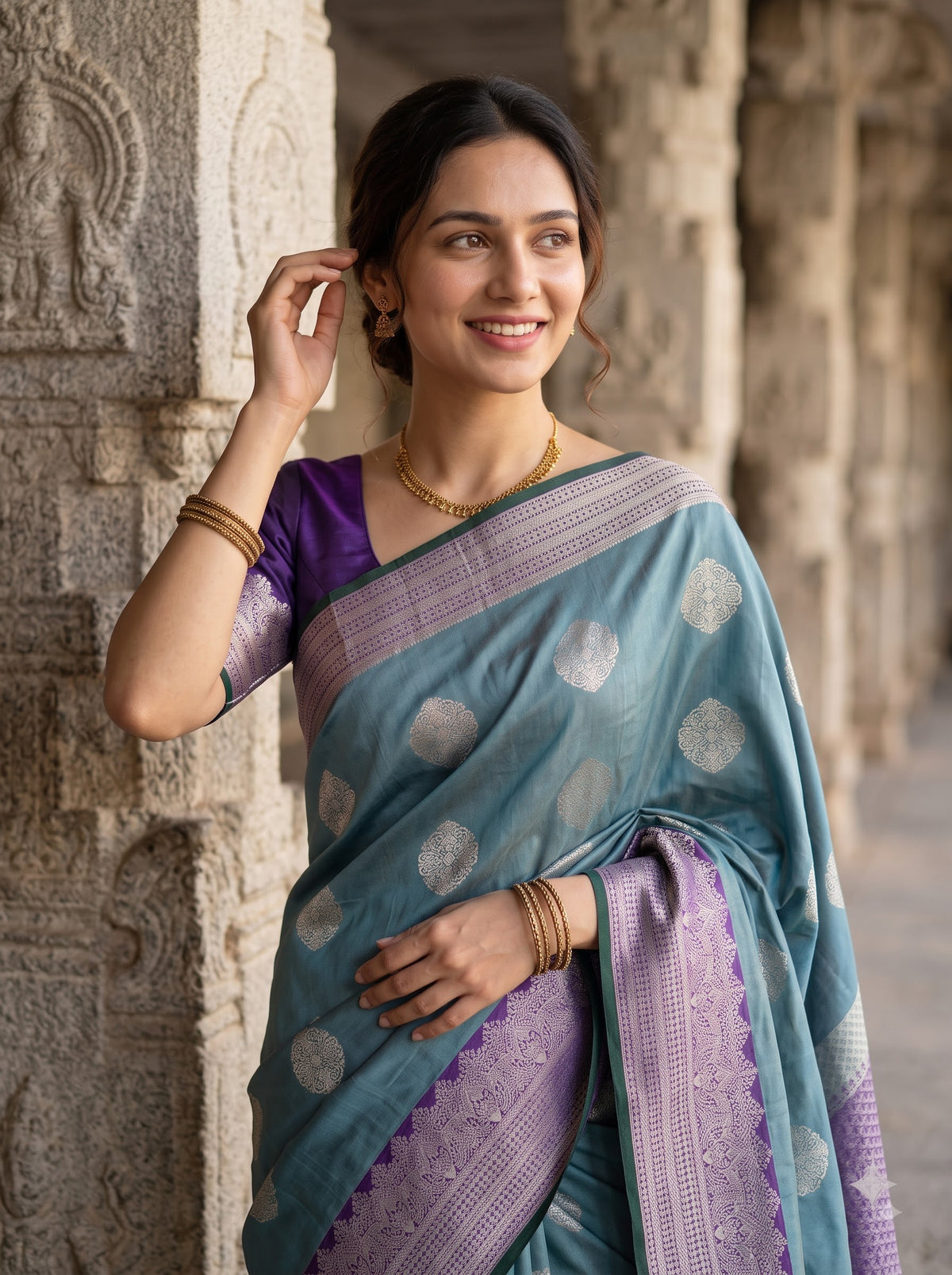 A woman wearing a teal and purple Banarasi saree with silver zari motifs, standing in a stone corridor.