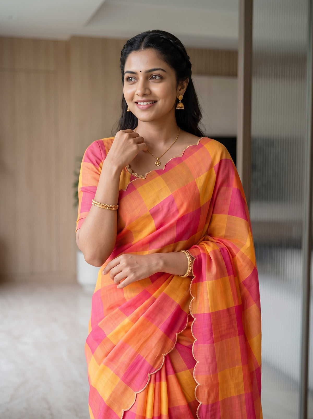 A woman wearing a bright orange and pink checkered saree with a scalloped border, standing in a modern indoor setting.