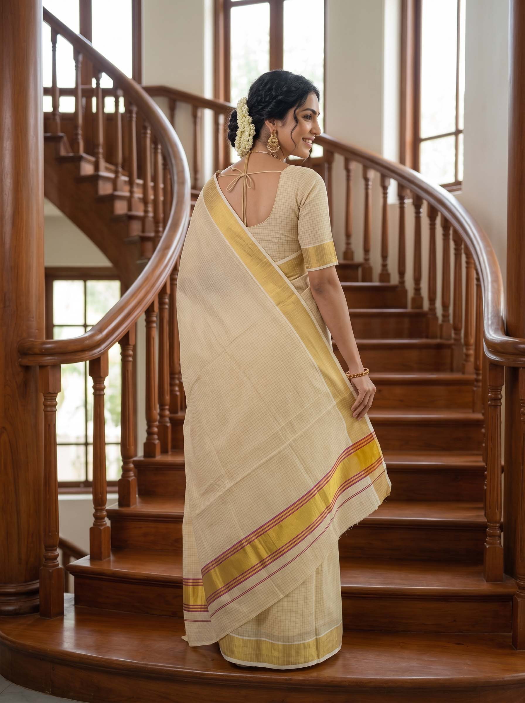 A woman wearing a cream-colored saree with yellow and red border stripes, standing on a wooden staircase in a home interior.