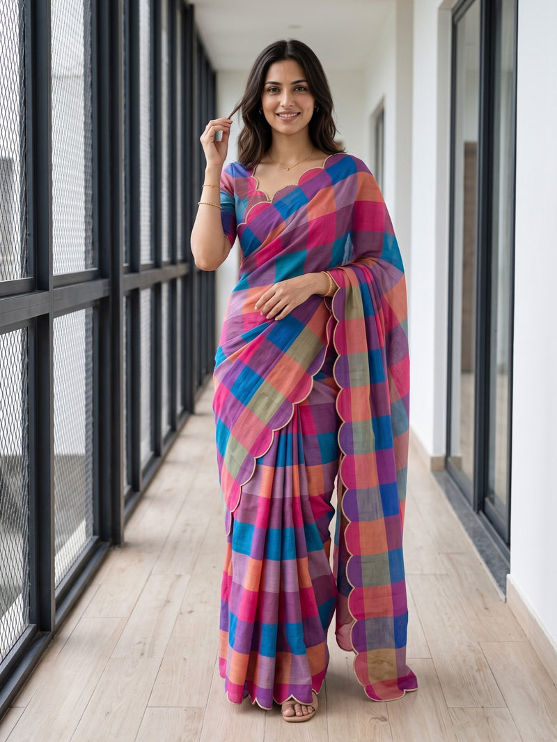 A woman wearing a vibrant multicolor checkered saree with pink, blue, orange, and purple tones, standing in a modern hallway with large windows.