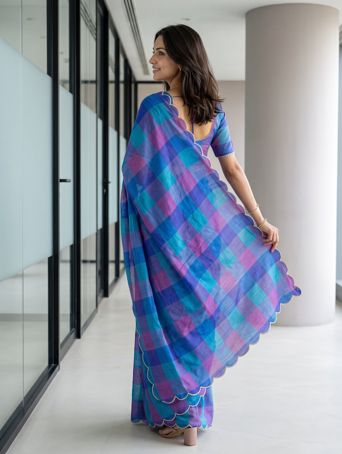 A woman wearing a blue and purple plaid saree with a matching blouse, standing in a modern indoor hallway with glass walls and a large white column.