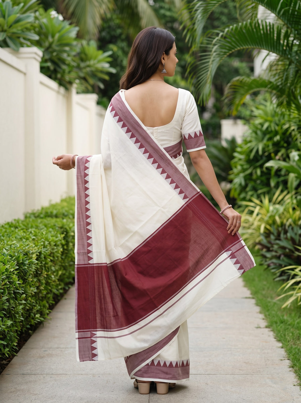 A woman wearing a white and maroon saree with geometric borders, photographed from behind as she stands on a garden path.