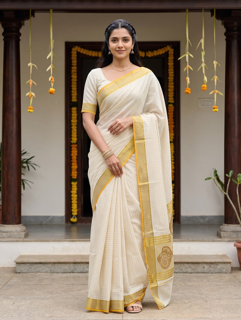 A woman stands in front of a traditional doorway wearing a cream-colored saree with a yellow border and pallu. The saree has a subtle grid pattern, and the background features wooden pillars, hanging yellow garlands, and a stone floor.