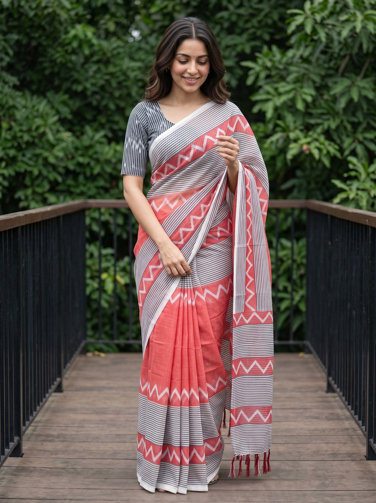 A woman wearing a coral pink and gray patterned saree with a matching gray printed blouse, standing on a wooden deck with green foliage in the background.