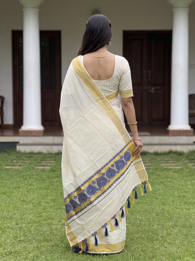 A woman stands outdoors on a lawn facing a building with white columns, wearing a cream-colored saree with yellow and blue border details and a matching yellow blouse.