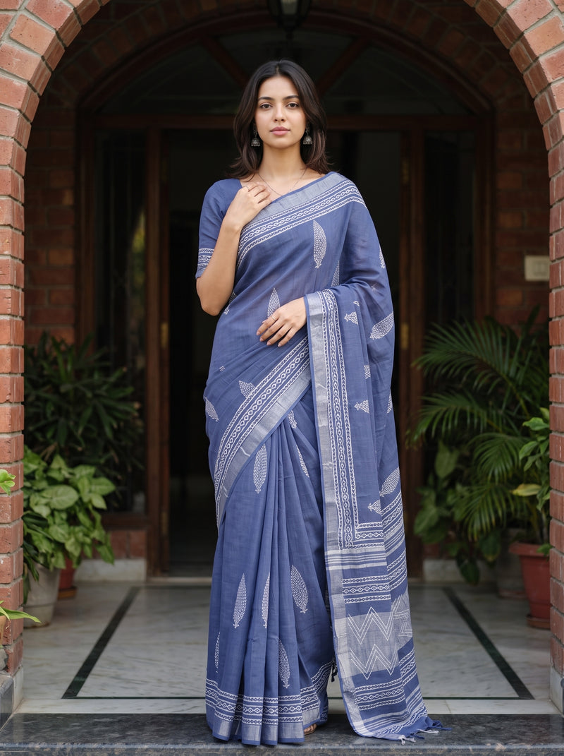 A woman wearing a slate blue saree with white printed motifs stands in front of a brick archway doorway with potted plants on either side.