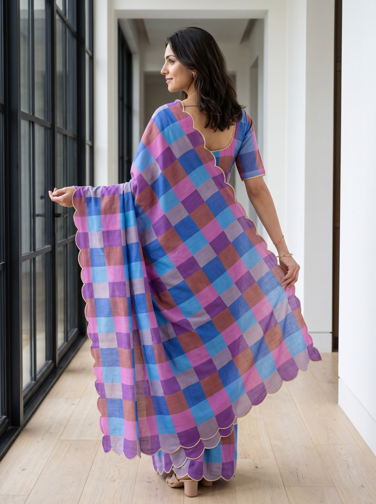 A woman wearing a purple, blue, and pink checkered saree with scalloped edges, standing in a hallway with light wood floors and white walls.