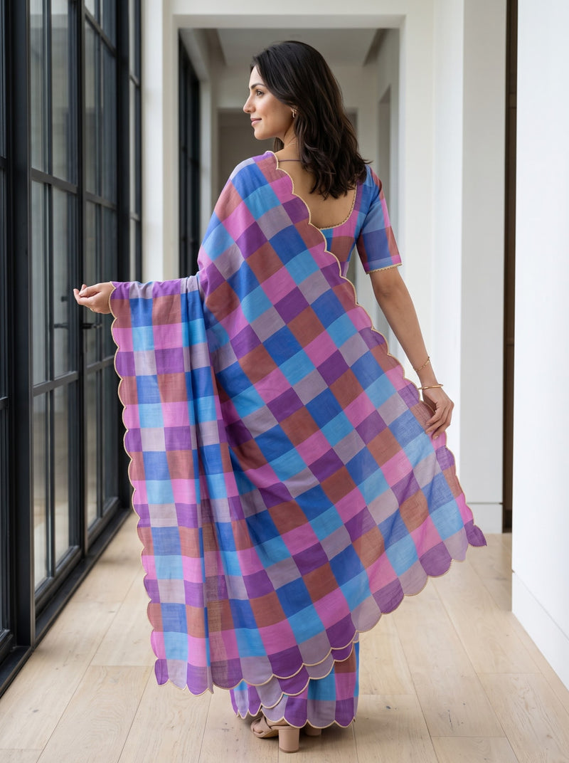 A woman wearing a purple, blue, and pink checkered saree with scalloped edges, standing in a hallway with light wood floors and white walls.