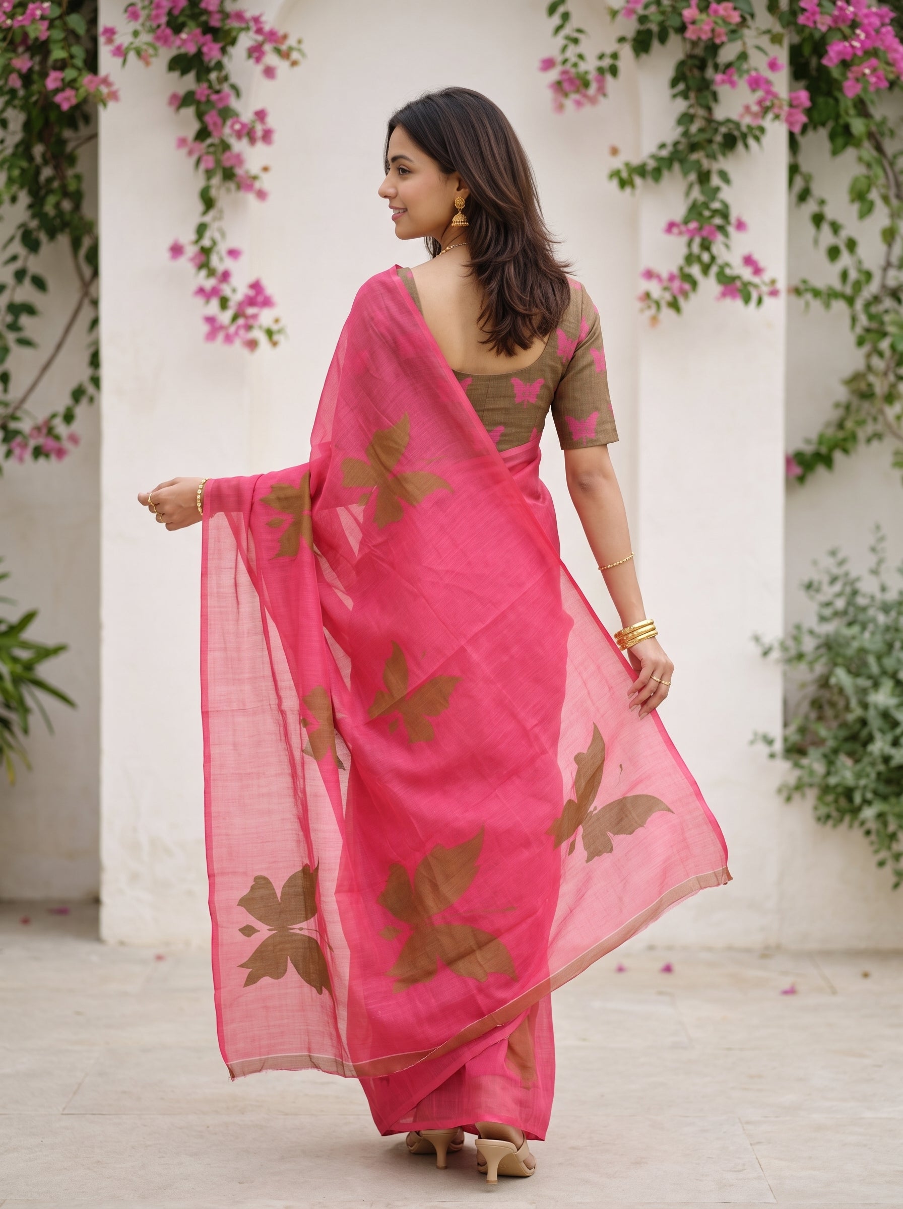 A woman wearing a bright pink saree with brown floral prints and a matching brown blouse, standing in front of a white wall with pink flowers.