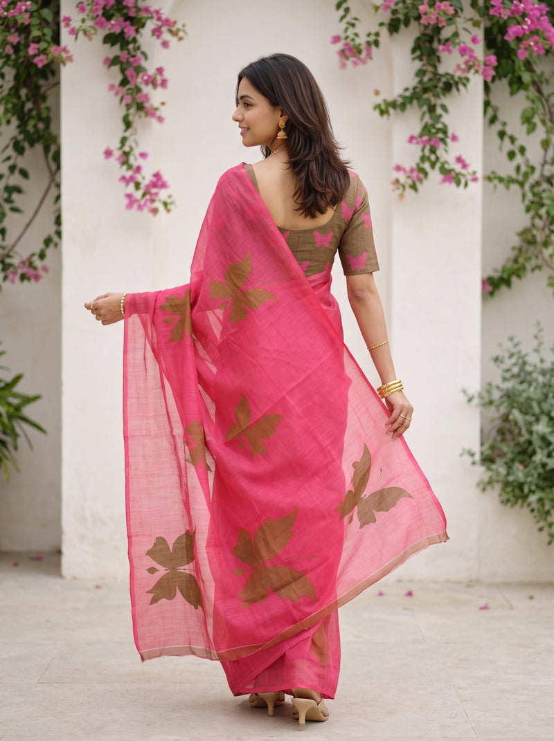 A woman wearing a bright pink saree with brown floral prints and a matching brown blouse, standing in front of a white wall with pink flowers.