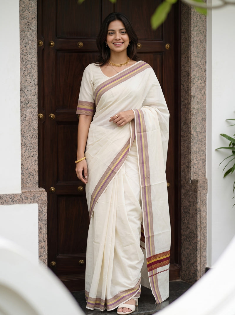 A woman wearing a cream-colored cotton saree with brown and gold striped borders, standing in front of a dark wooden door.