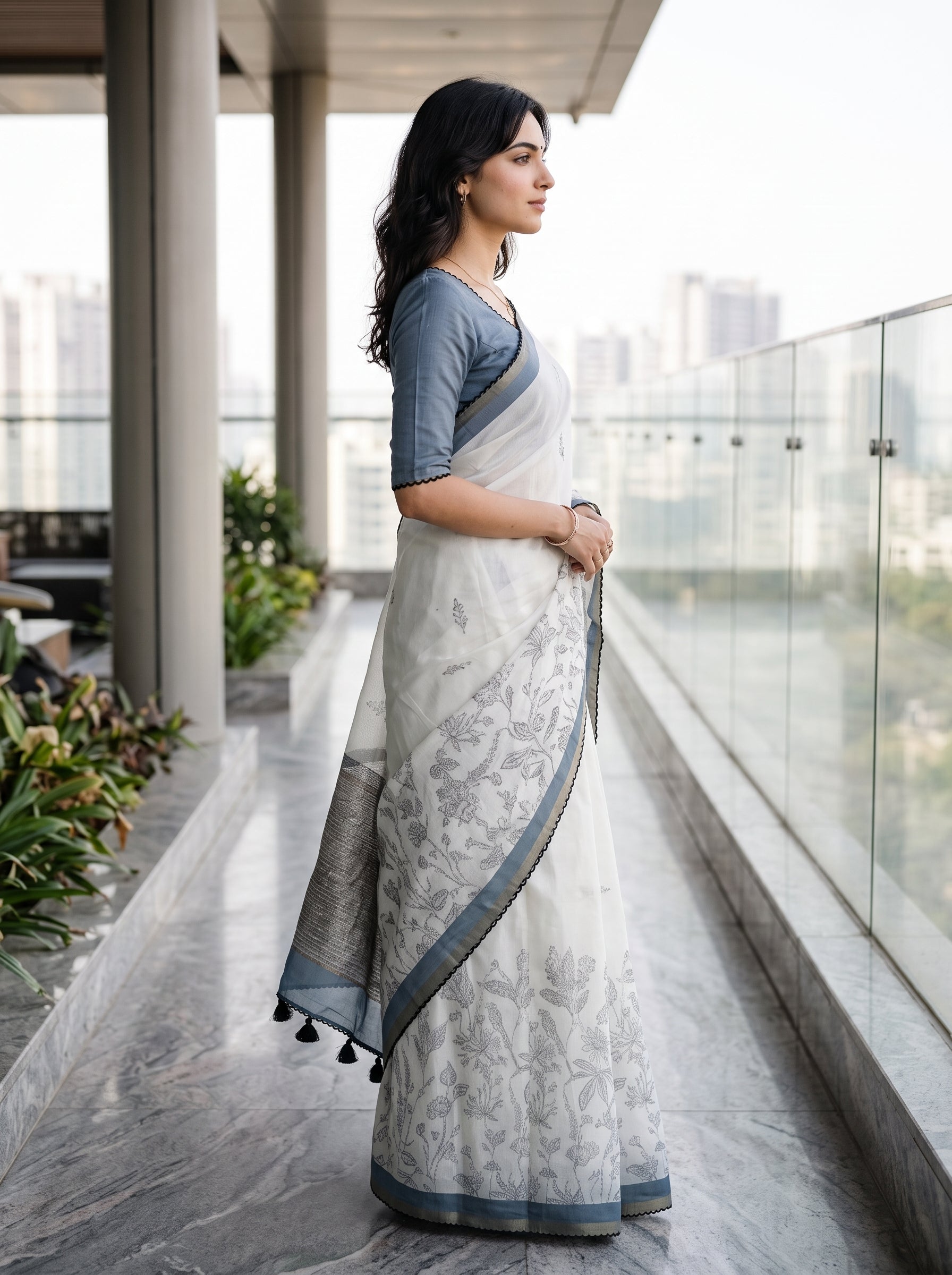 A woman wearing a white and gray floral-patterned saree with a slate blue blouse, standing on a modern glass-rail balcony overlooking a city skyline.