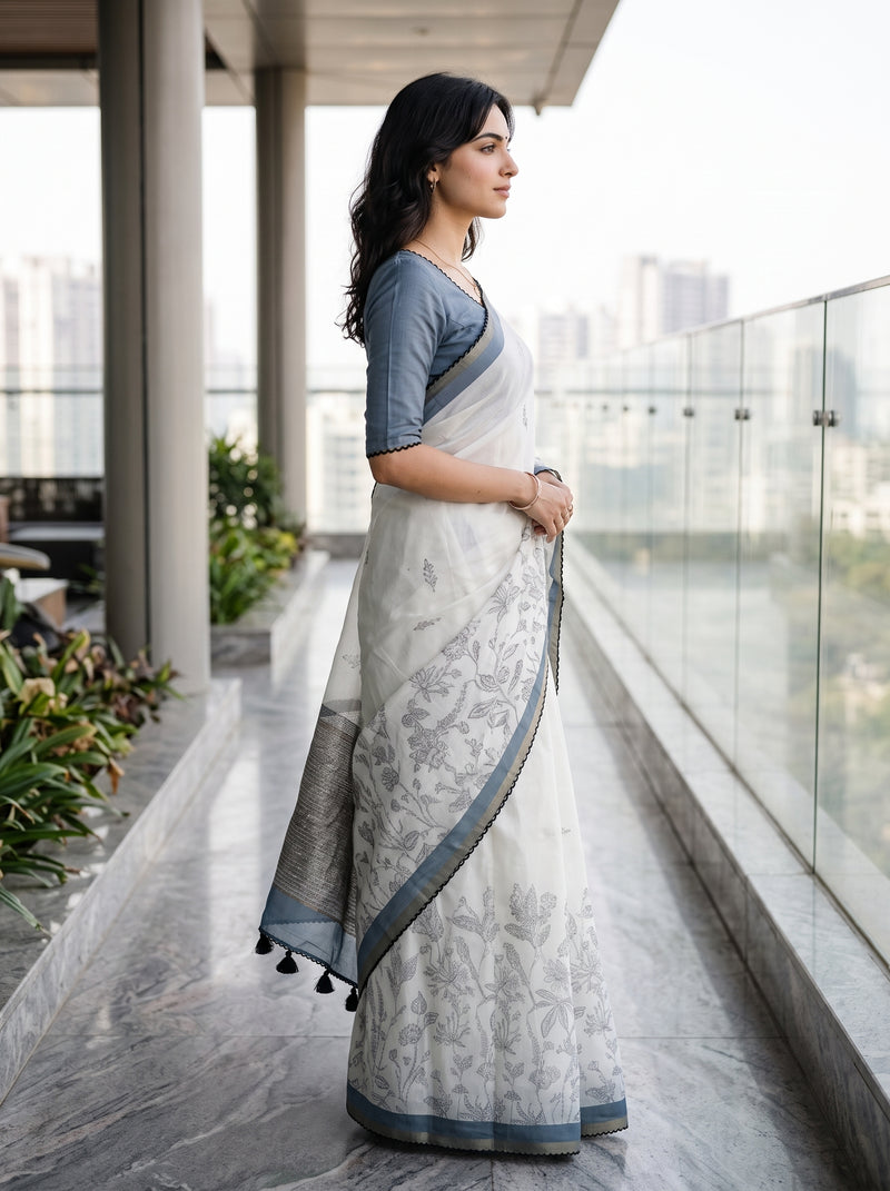 A woman wearing a white and gray floral-patterned saree with a slate blue blouse, standing on a modern glass-rail balcony overlooking a city skyline.