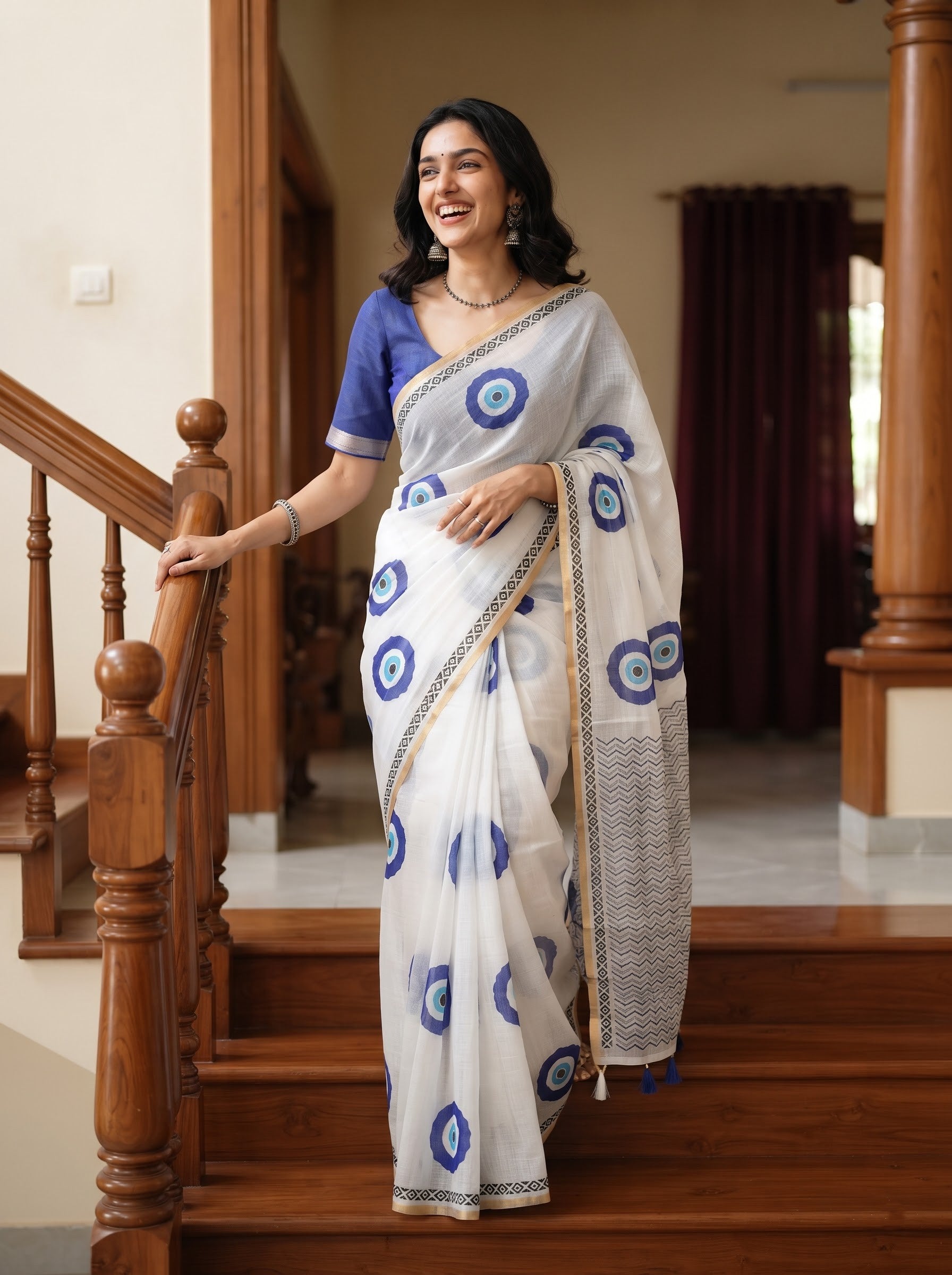 A woman wearing a white saree with blue circular motifs and a blue blouse, standing on wooden stairs in an indoor setting.