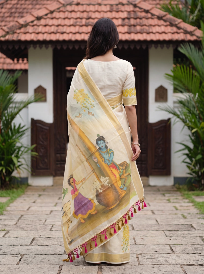 A woman viewed from behind wearing a cream-colored saree with a detailed hand-painted illustration of Lord Krishna and a decorative border with pink tassels, standing in front of a traditional house with a tiled roof.