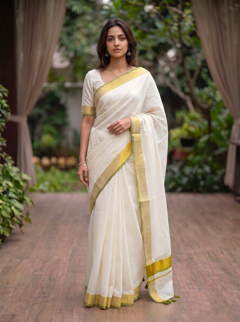 A woman wearing a white cotton saree with a yellow-gold border and pallu, standing in a garden courtyard setting.