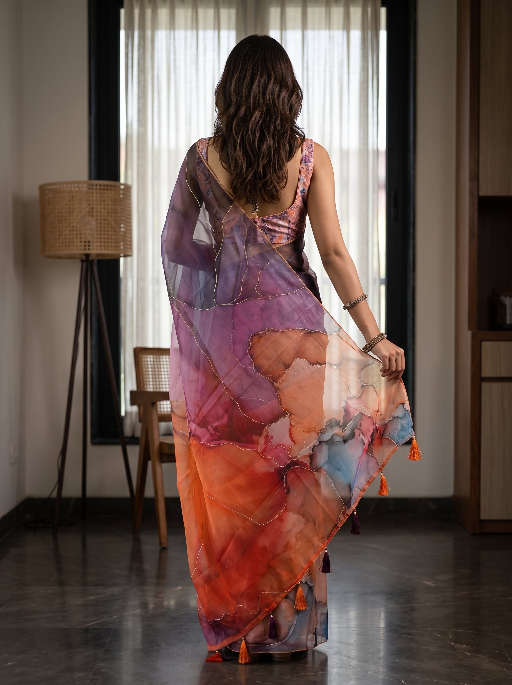 A woman viewed from behind wearing a sheer, multicolored printed saree with orange, purple, pink, and blue watercolor-like patterns and small orange tassels, standing in a modern indoor room.