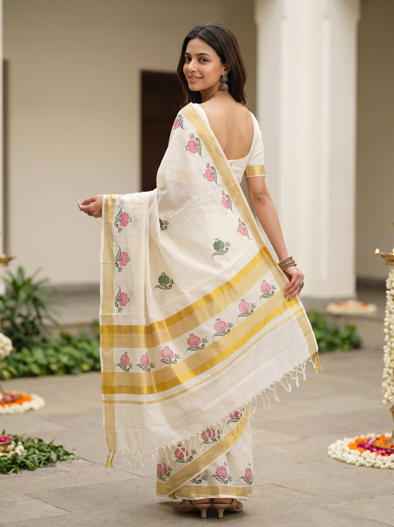 A woman wearing a white cotton saree with pink floral prints and yellow-gold borders, standing in a courtyard setting.