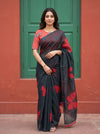A woman wearing a black and red floral printed saree with a red sleeveless blouse, standing in front of a green door and terracotta wall.