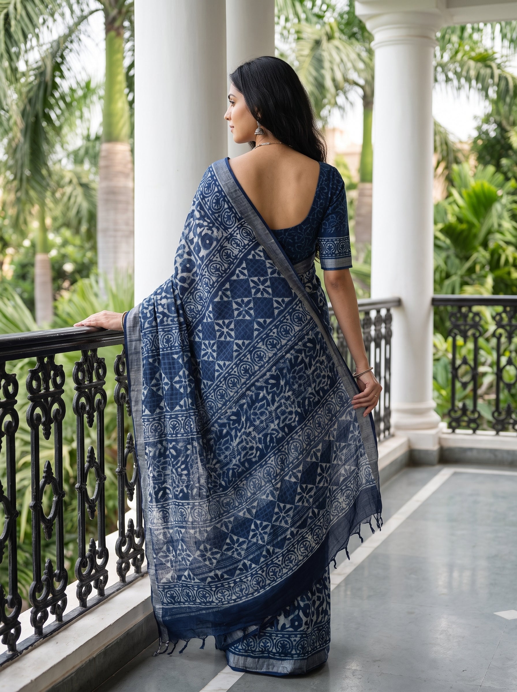 A woman wearing a navy blue and white printed saree stands on a balcony with white pillars and green foliage in the background, shown from the back.