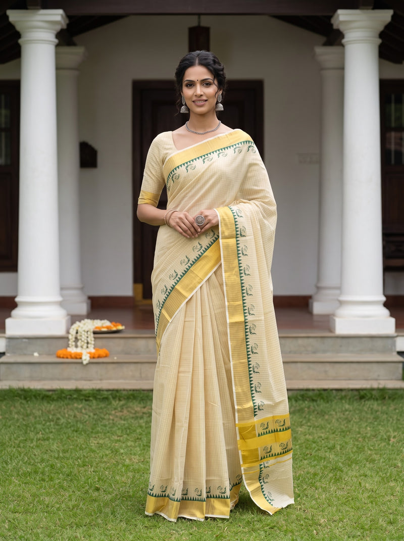 A woman stands outdoors in front of a house with white columns, wearing a light beige saree with yellow and green border details and a matching blouse.