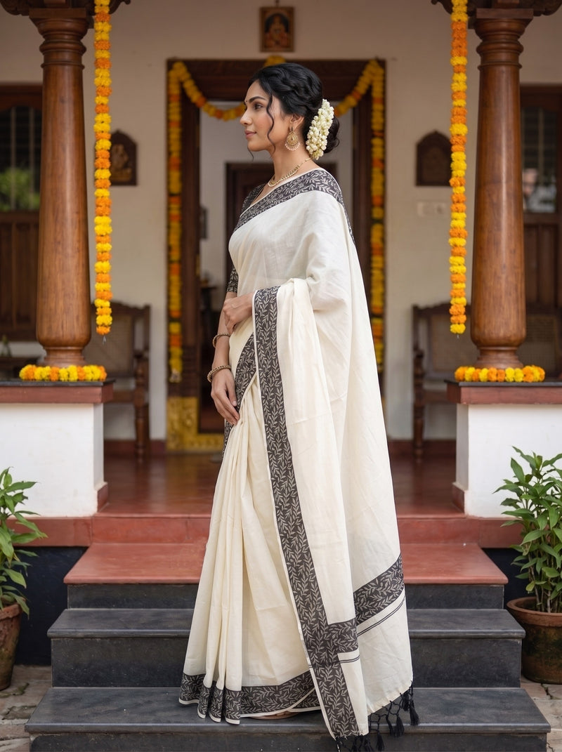 A woman wearing a cream-colored cotton saree with black border patterns, standing on steps in front of a traditional house decorated with yellow marigold garlands.