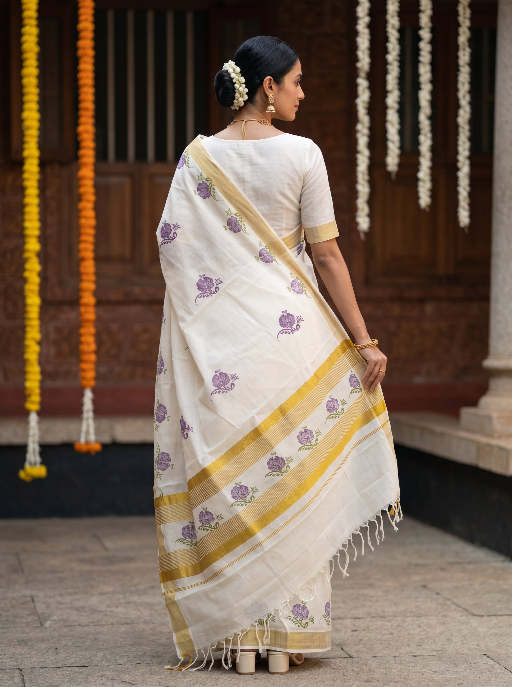 A woman wearing a white saree with purple floral prints and gold stripes, standing in front of a traditional wooden building with hanging marigold garlands.
