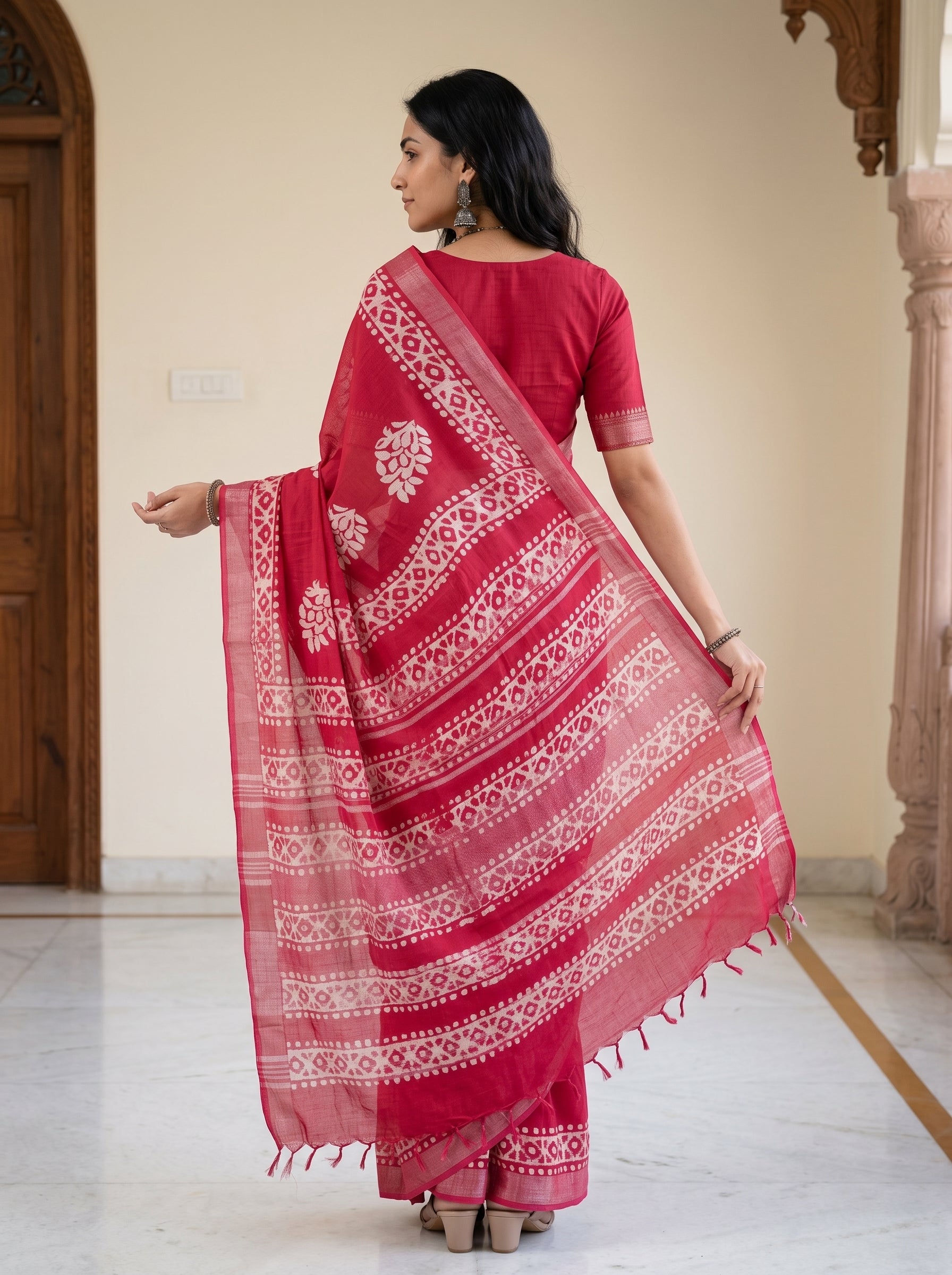 A woman wearing a red salwar suit with a matching red and white patterned dupatta, photographed from behind in an indoor architectural setting with cream walls and wooden columns.