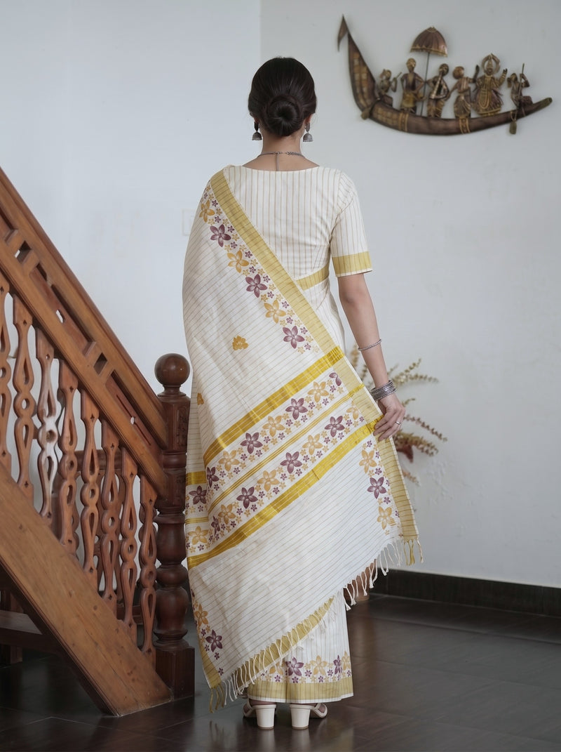 A woman viewed from behind wearing a cream-colored cotton saree with yellow and purple floral border patterns, standing in an indoor setting next to a wooden staircase.