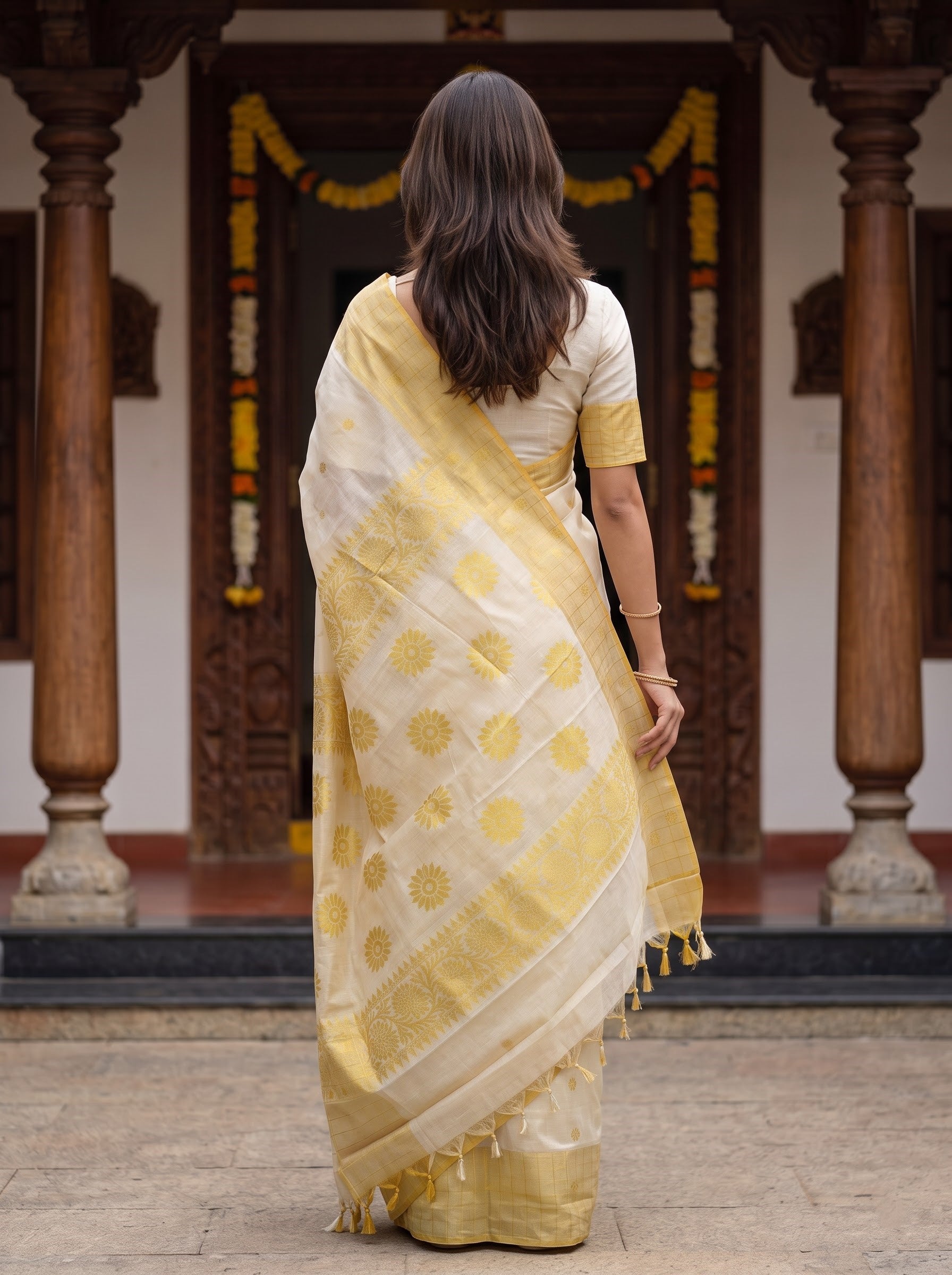 A woman viewed from behind wearing a yellow and white saree with gold motifs, standing in front of a traditional carved wooden doorway with pillars.