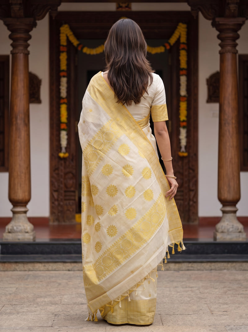 A woman viewed from behind wearing a yellow and white saree with gold motifs, standing in front of a traditional carved wooden doorway with pillars.