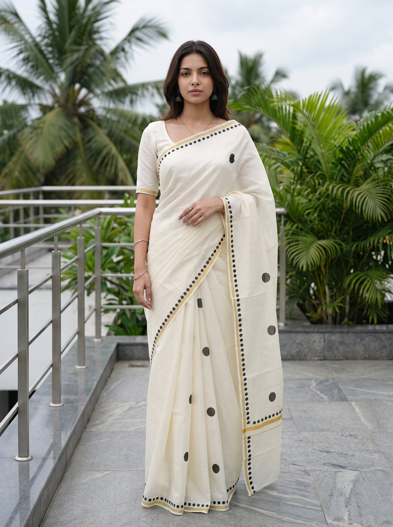 A woman wearing a cream-colored cotton saree with black polka dots and gold border detailing, standing on an outdoor balcony with palm trees in the background.