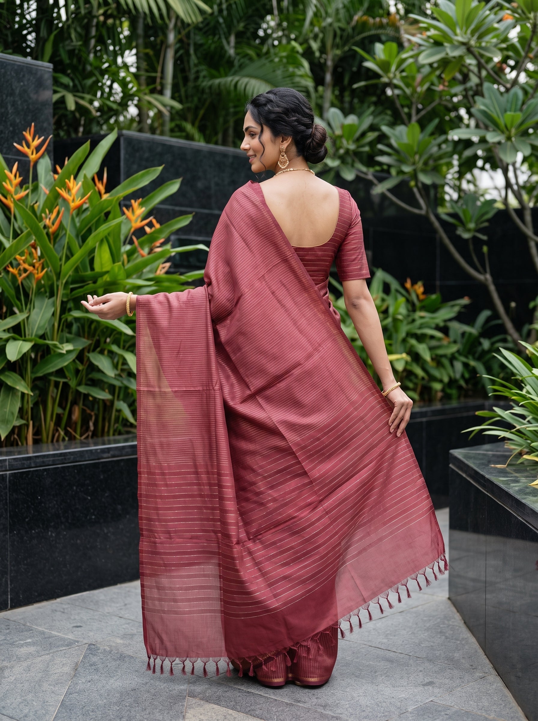 A woman wearing a dusty rose terracotta-colored cotton saree with a matching blouse, standing outdoors in a garden setting with green foliage and black stone planters.