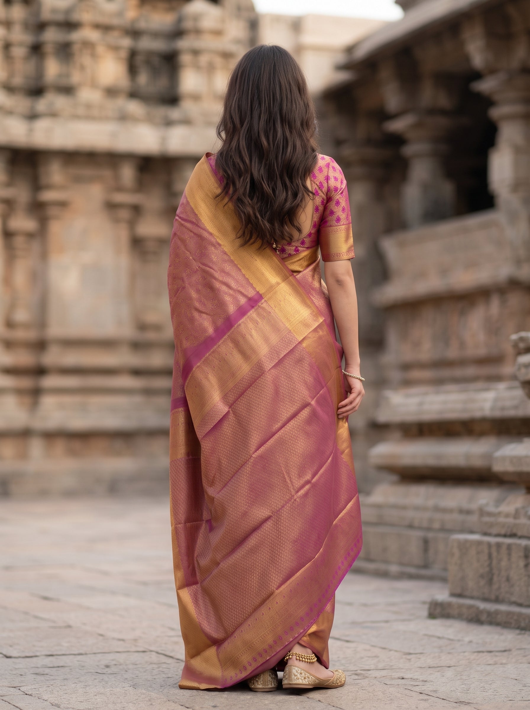 A woman viewed from behind wearing a traditional Indian saree with a pink and gold patterned design, standing in front of a stone temple structure.