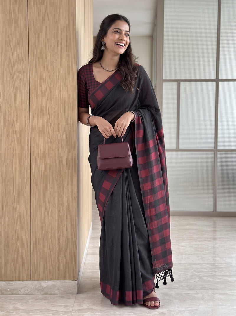 A woman wearing a black and red checkered saree with a matching checkered blouse, holding a small burgundy handbag, standing in a modern indoor setting with light wood paneling.