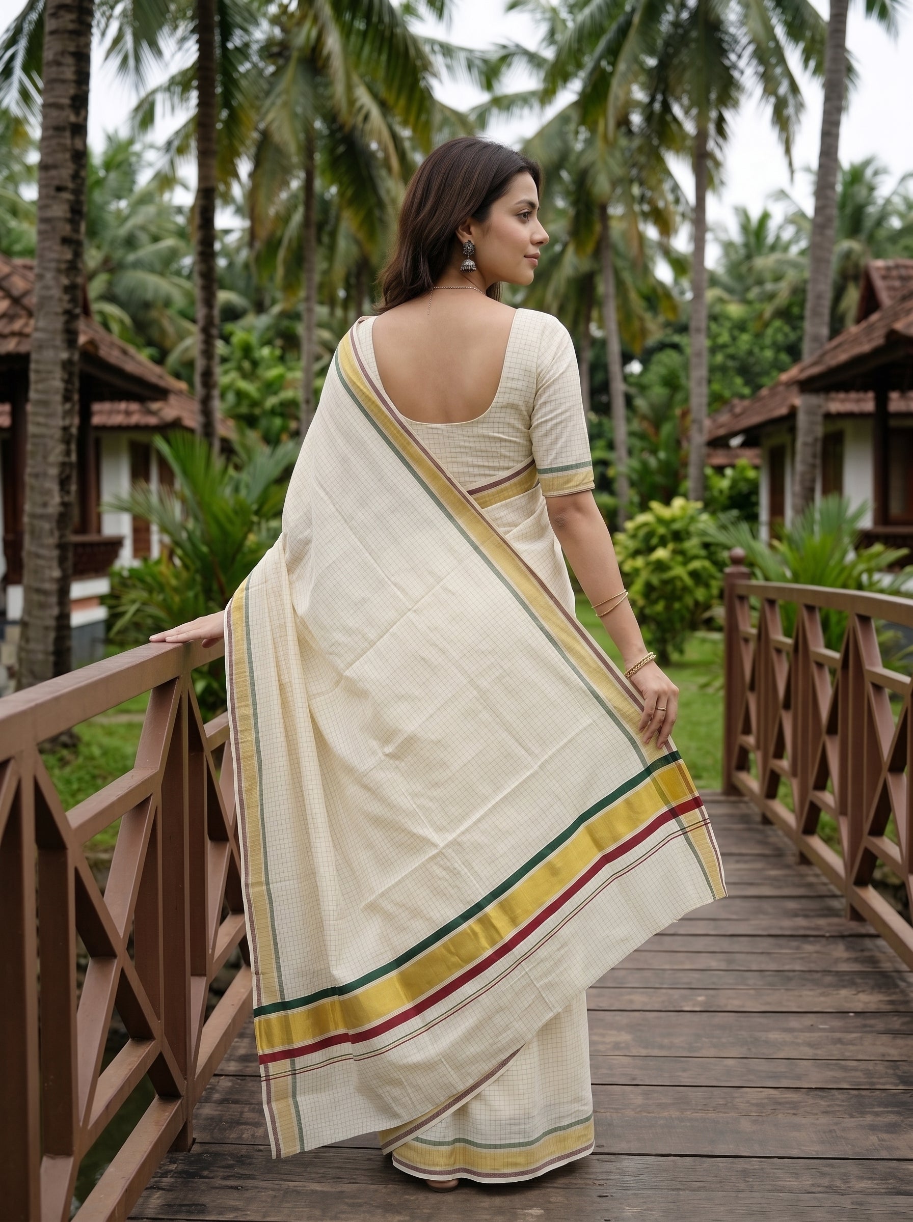 A woman wearing a cream-colored cotton saree with yellow, green, and red border stripes, standing on a wooden bridge in a tropical resort setting with palm trees.