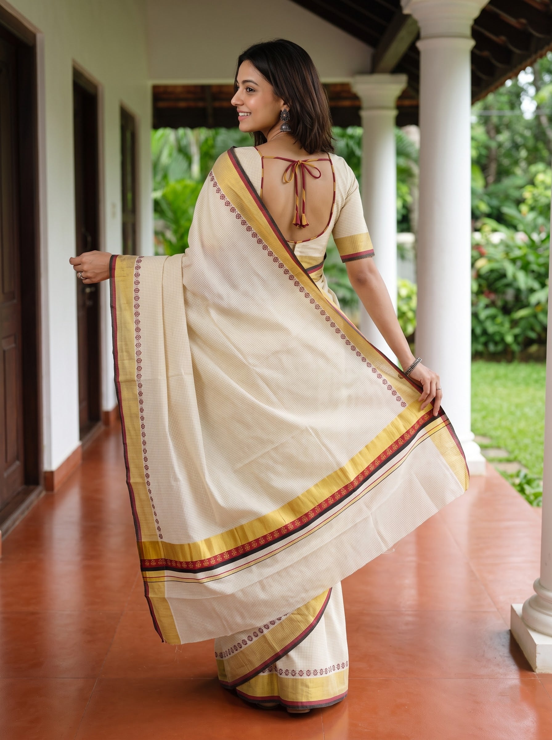 A woman wearing a cream-colored cotton saree with yellow, red, and blue border stripes, standing on a tiled porch with white pillars and green garden foliage in the background.
