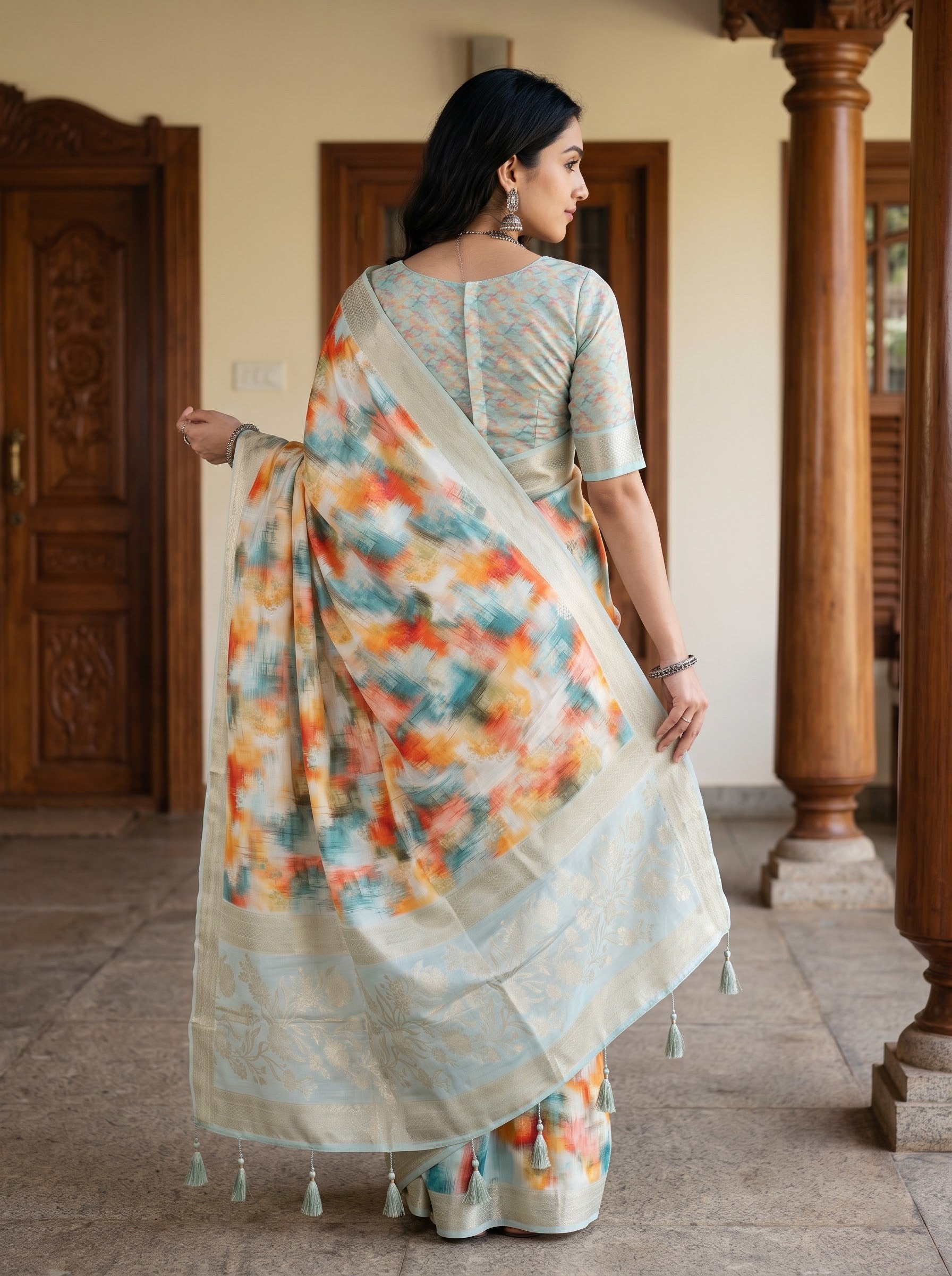 A woman wearing a light sage green saree with a colorful tie-dye floral pattern in orange, yellow, and blue, standing in a traditional courtyard with wooden pillars.
