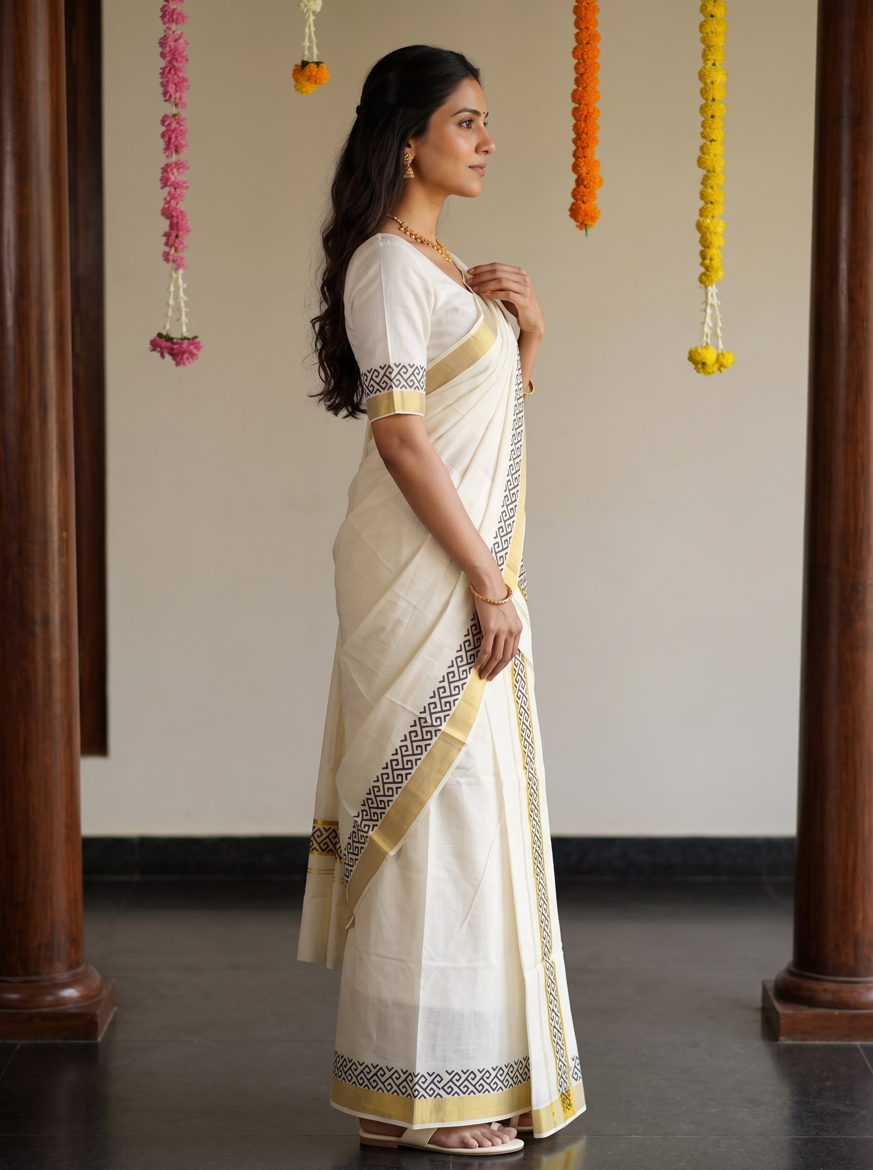 A woman wearing a traditional Indian saree in cream and gold, standing in profile in an indoor setting with decorative garlands hanging in the background.
